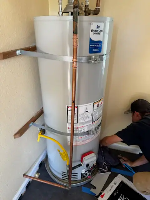A man kneels next to a tall, white Bradford White water heater installed in a corner. The unit is secured to the wall with metal straps and connected to pipes at the top and side. Tools and a cloth are on the floor nearby.