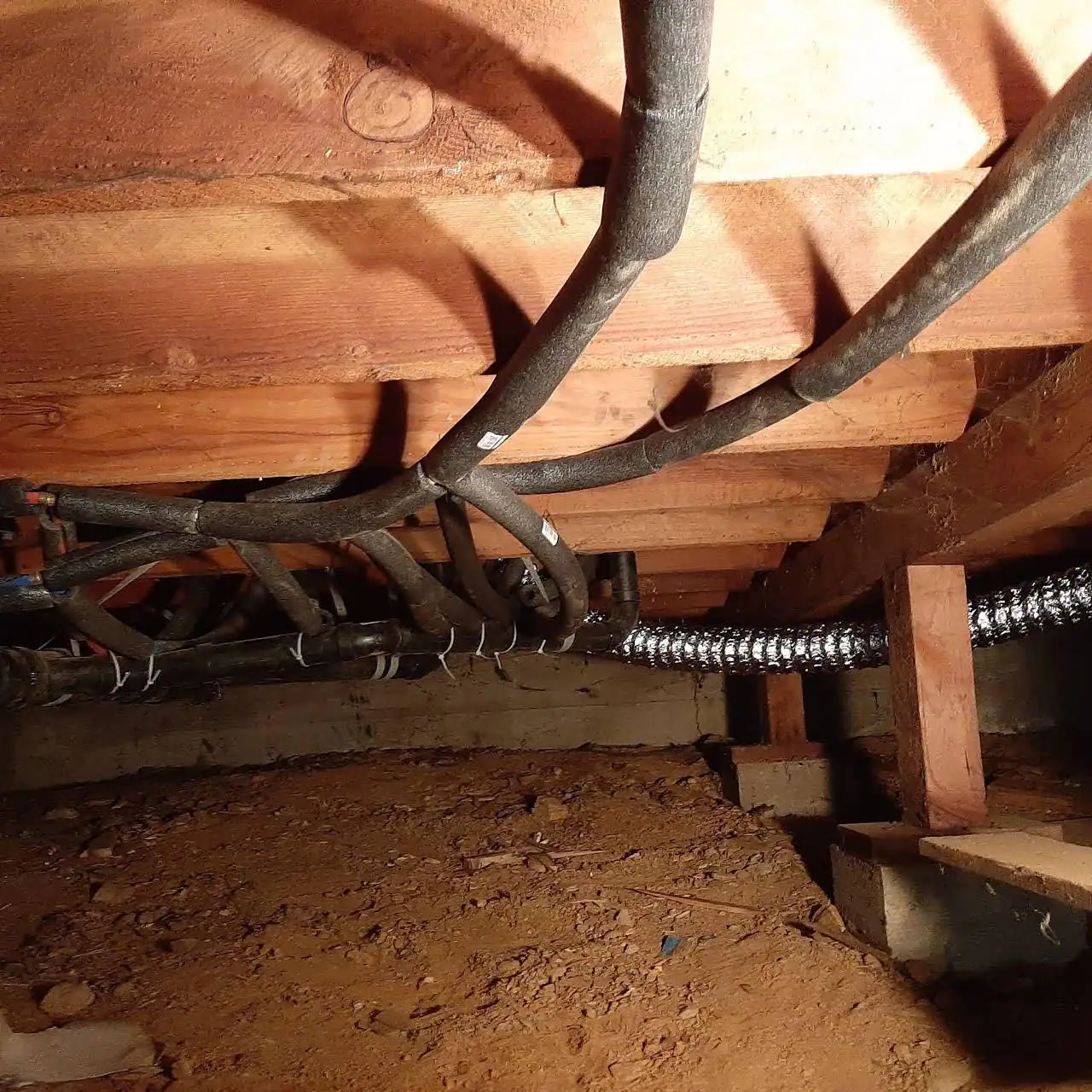 A crawl space with an exposed dirt floor, wooden ceiling beams, insulated pipes, and a flexible metal duct running along the beams—an unfinished area often accessed by a plumber El Dorado County, CA for utility work or repairs.