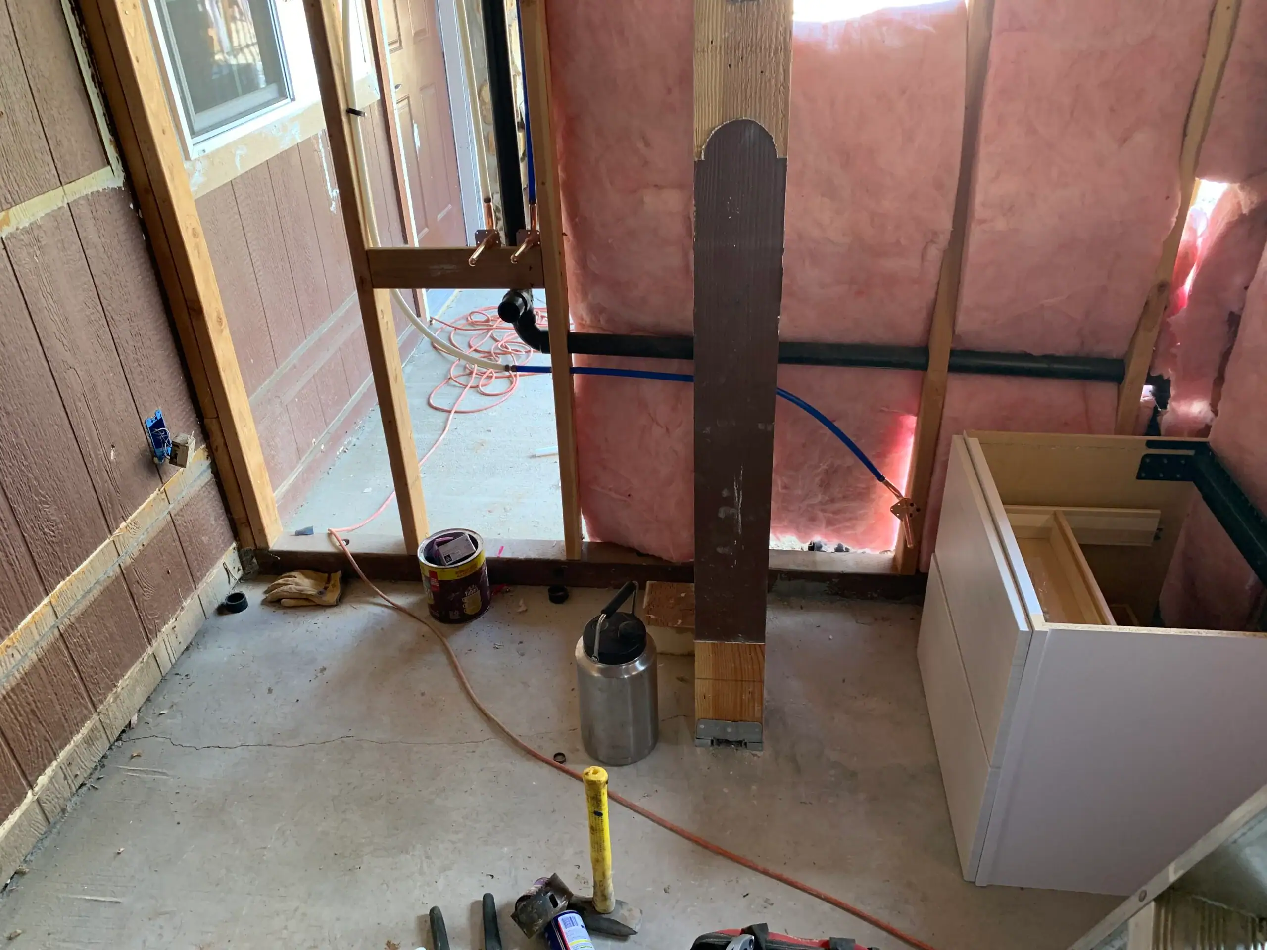 Partially finished room under construction with exposed wall framing, pink insulation, plumbing pipes ready for a water heater replacement El Dorado County, electrical wiring, cabinet, tools, tape measure, and a can of paint on the concrete floor.