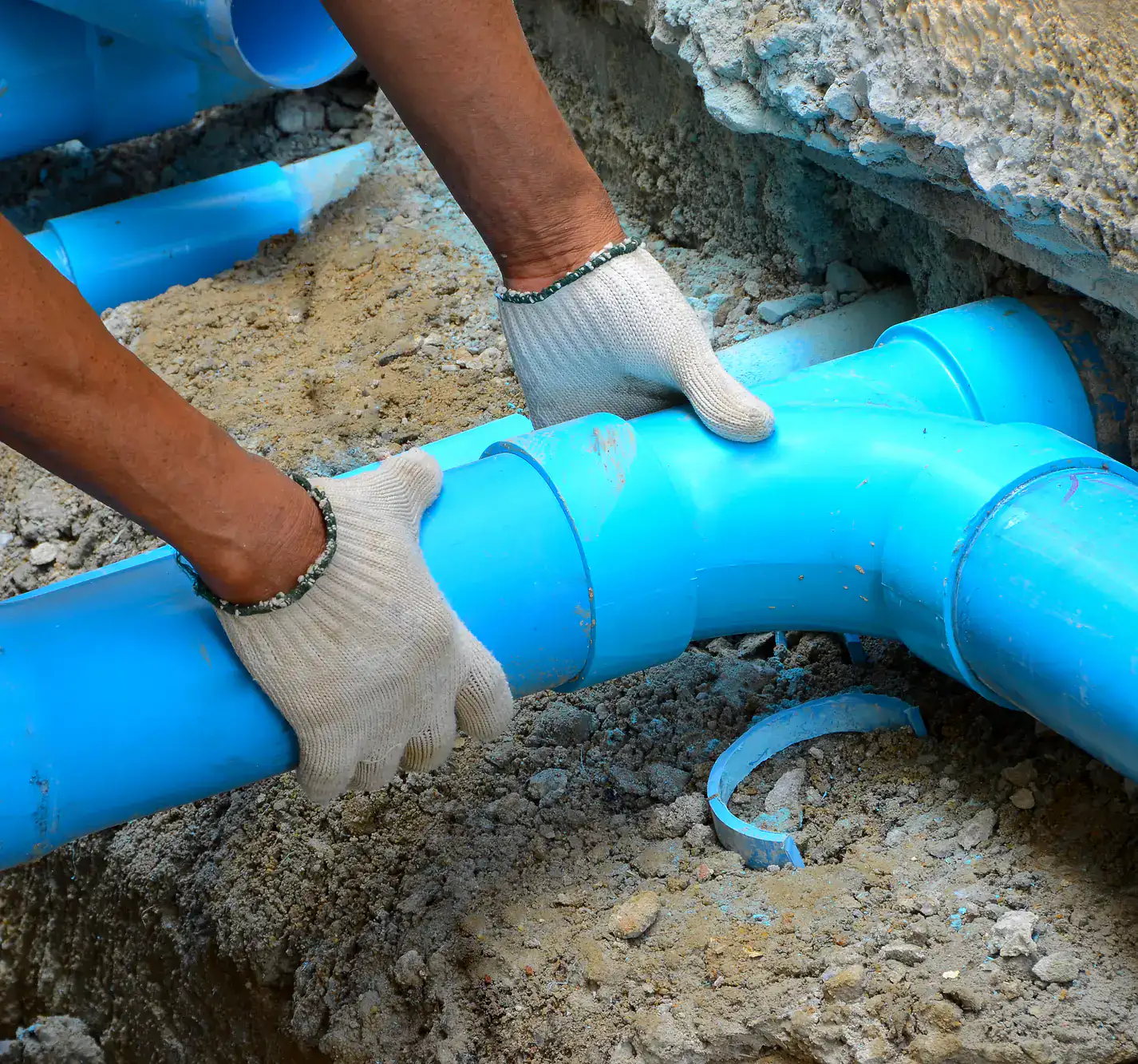 A plumber in El Dorado County, CA, wearing white gloves, connects bright blue PVC pipes in a dirt-filled trench—likely working on an underground plumbing installation or repair.
