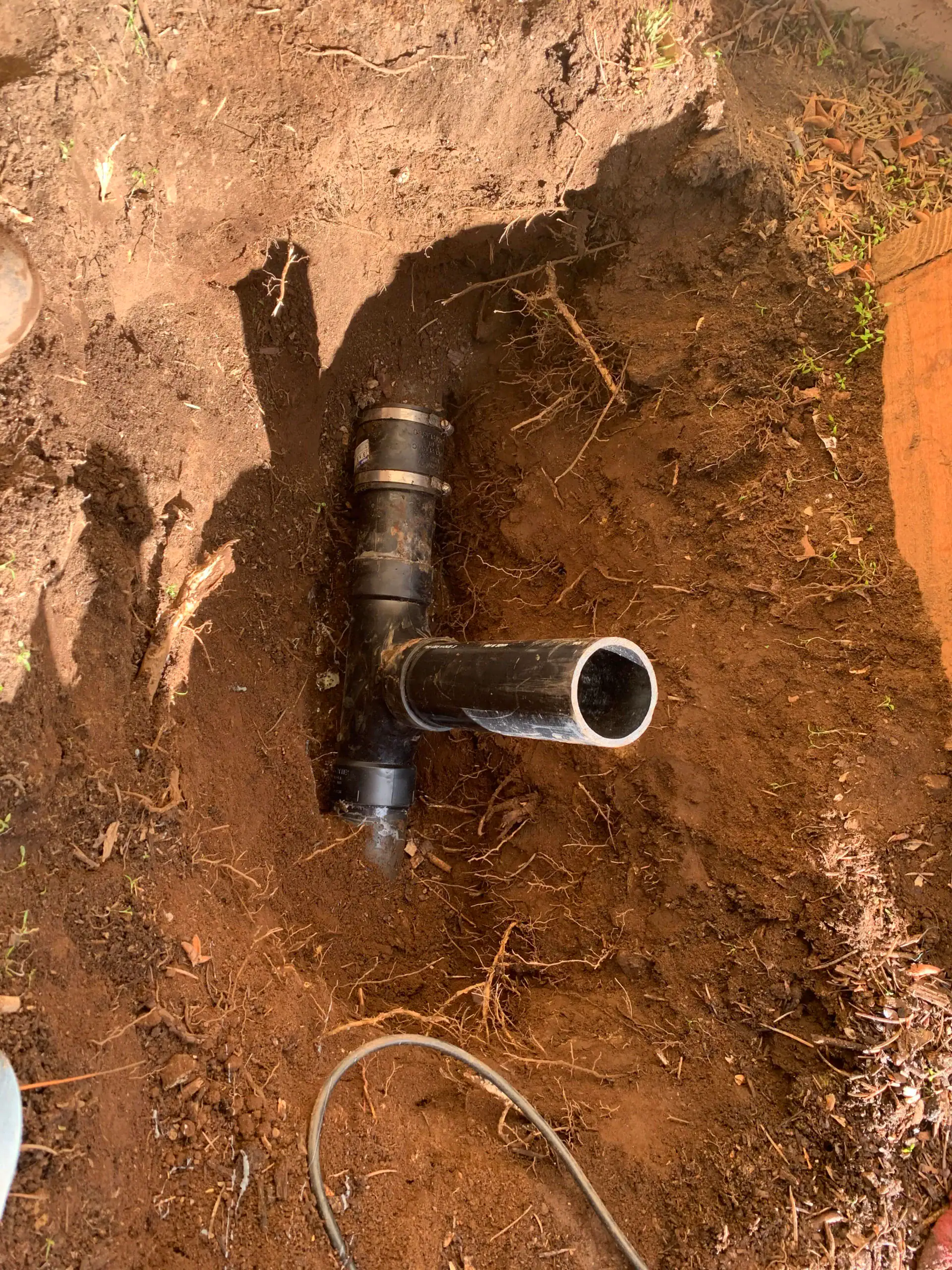 A black PVC T-joint pipe is partially buried in soil, surrounded by loose dirt and a nearby cable, as sunlight casts shadows—possibly part of a plumber El Dorado County project or water heater replacement El Dorado County, CA.