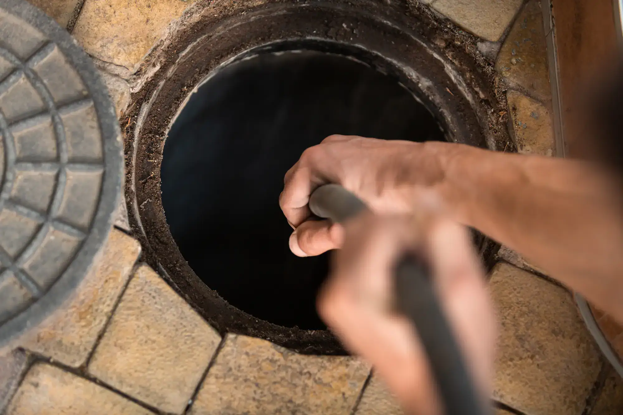 A plumber El Dorado County is shown holding a tool while cleaning or inspecting the inside of an open, round manhole surrounded by tan, stone tiles.