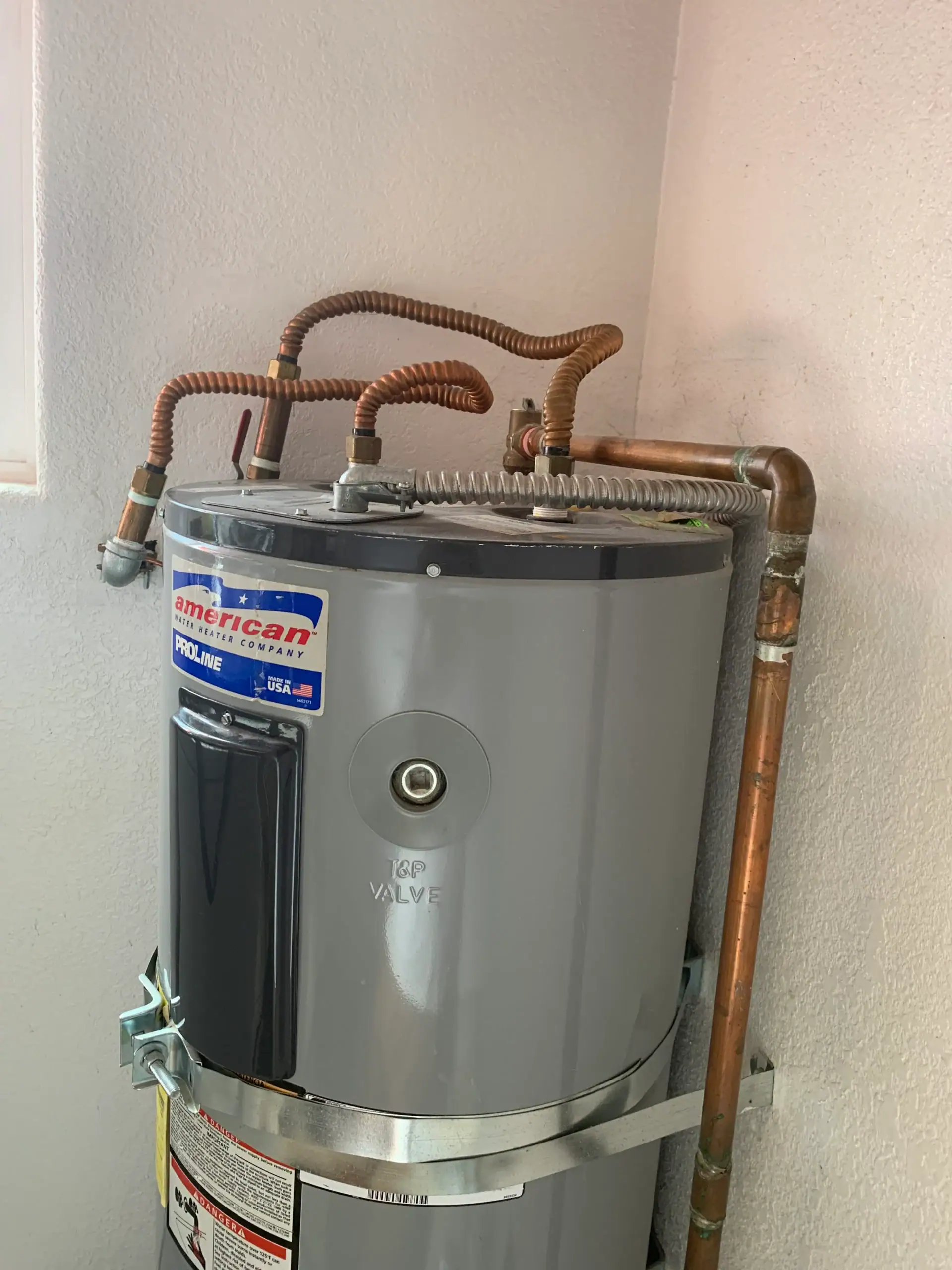 A gray American ProLine water heater with multiple copper pipes connected at the top, installed in a corner against white walls. For water heater replacement El Dorado County, CA residents can trust local professionals for expert installation.