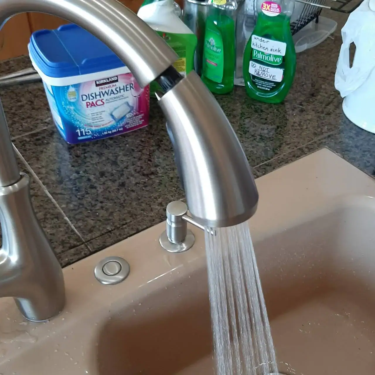 A kitchen faucet with water running into a beige sink. On the granite countertop are a blue dishwasher pacs container, green dish soap bottles, and other kitchen items—a tidy space for any plumber El Dorado County, CA would appreciate.