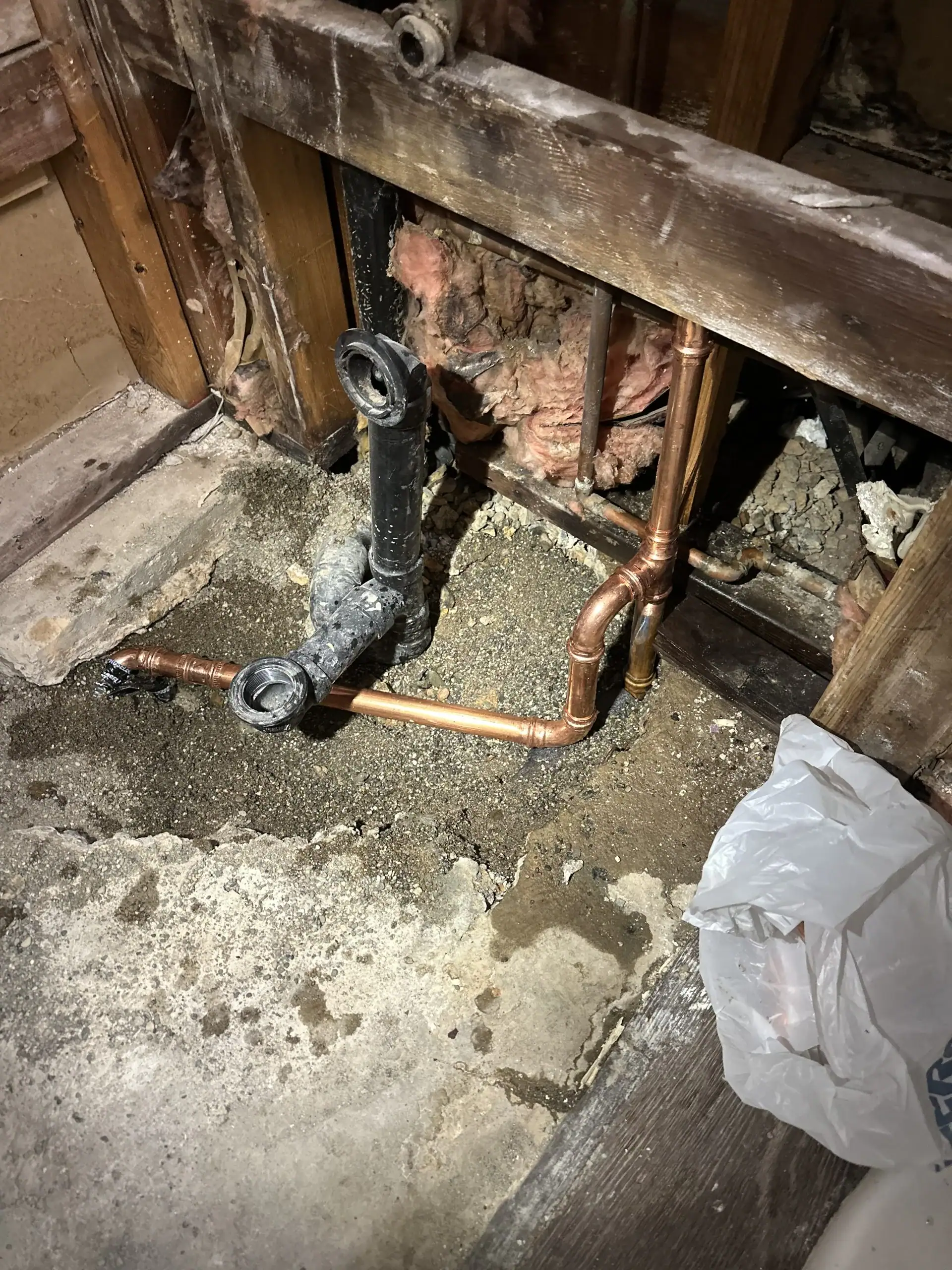 Exposed plumbing pipes, insulation, and framing are visible in a wall under repair. A plumber El Dorado County might uncover copper pipes and a black drainpipe surrounded by dirt and concrete during a CA water heater replacement. A plastic bag sits nearby.
