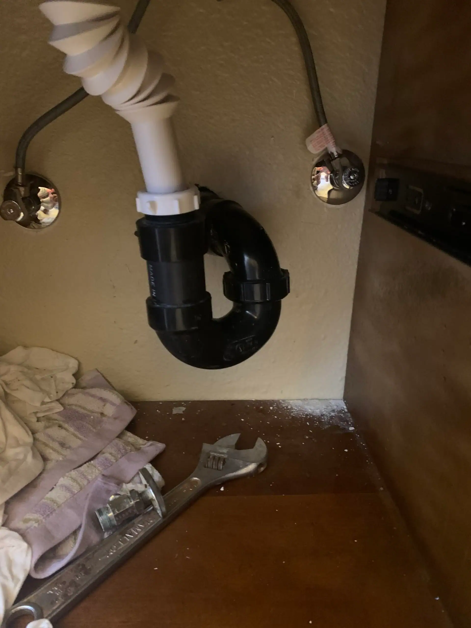 Under-sink plumbing with a black P-trap, white drainpipe, exposed water lines, a wrench, a glass cup, and cloth—plus some white residue on the cabinet floor—ready for a plumber El Dorado County, CA to inspect.