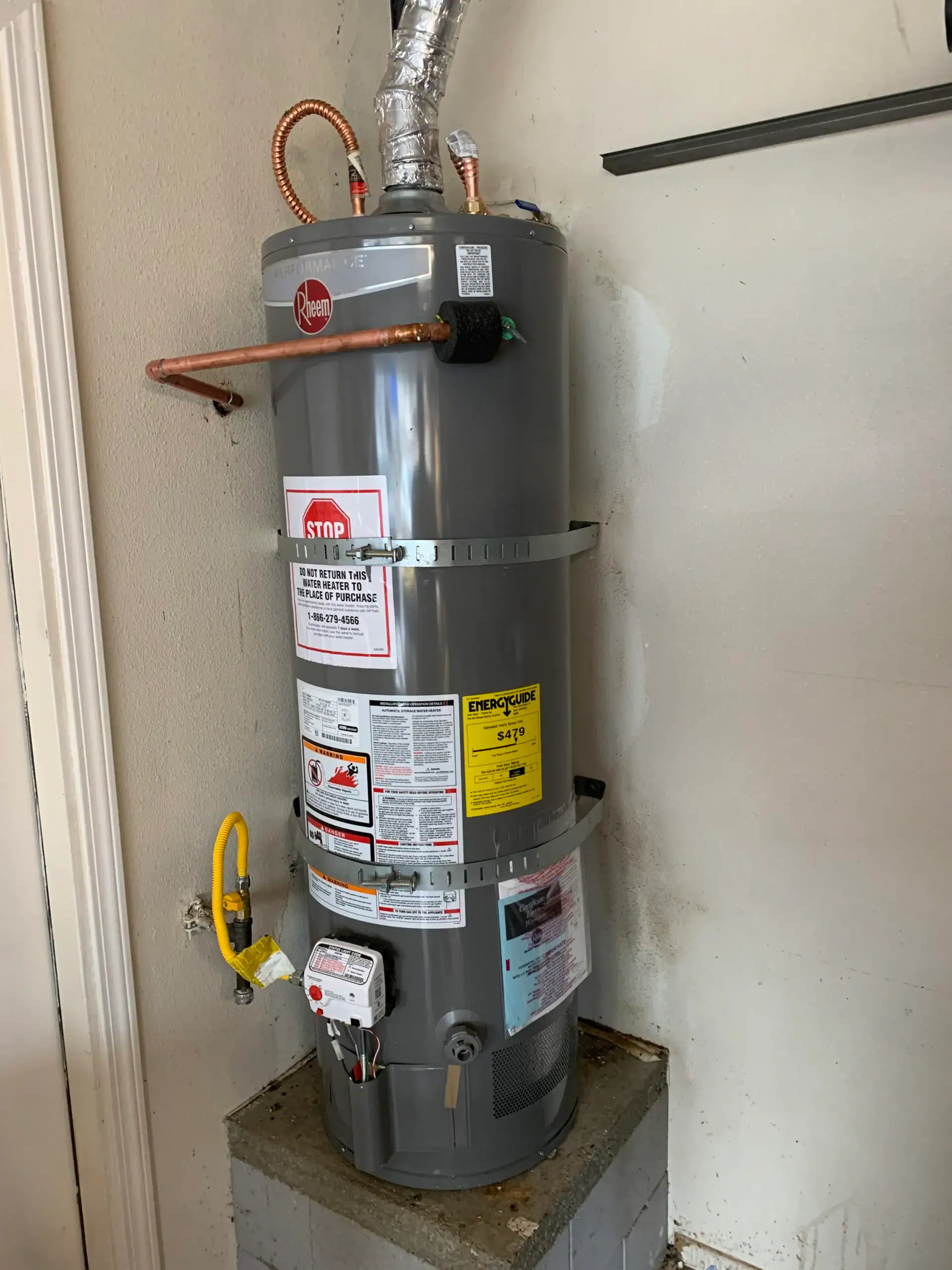A gray Rheem water heater is installed against a wall in a CA garage, secured with metal straps. It features warning labels, a yellow gas line, and connected pipes above and on the side—ready for expert plumber El Dorado County service.