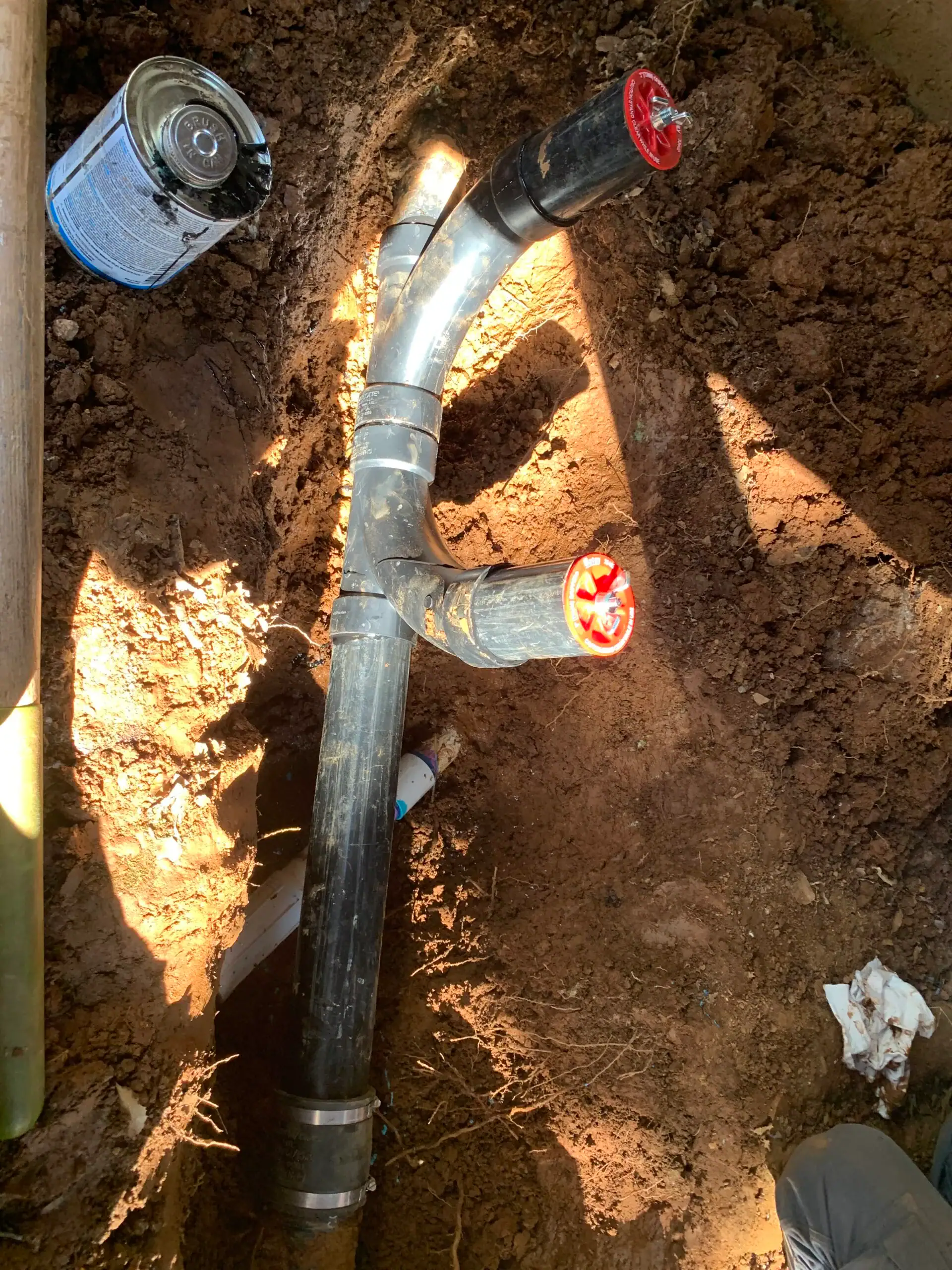 A black PVC pipe with a Y-junction, often seen in water heater replacement El Dorado County, CA projects, is partially buried in dirt. Both branch ends are capped in red while an open can of PVC cement sits nearby in the sunlight.