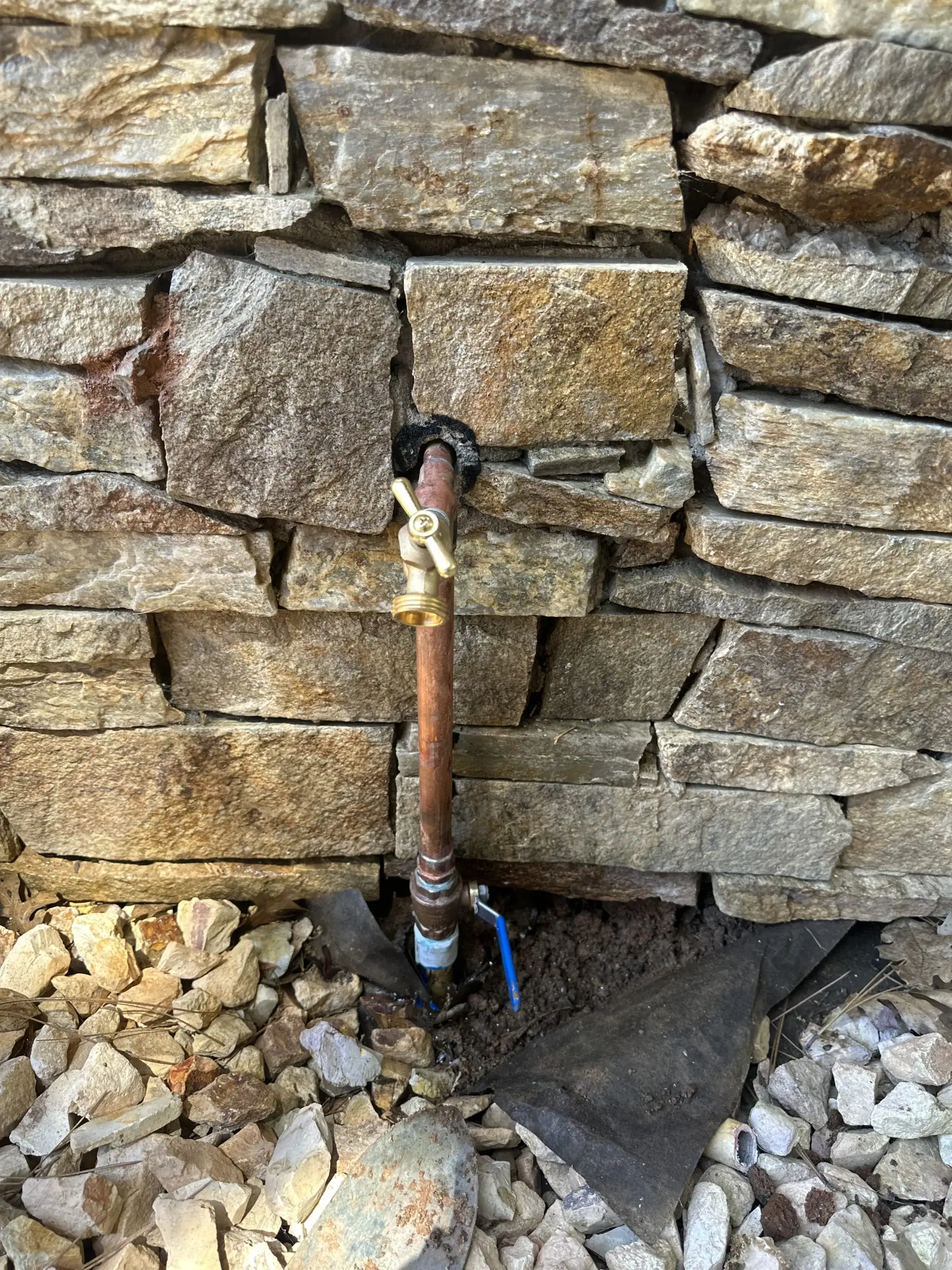Outdoor brass water tap attached to a copper pipe and stone wall, its base ringed by rocks and soil. A blue valve and black material sit near the ground—a classic setup often serviced by a plumber El Dorado County residents can trust.