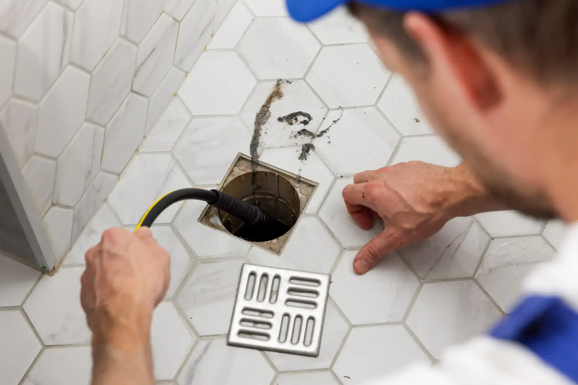 A plumber El Dorado County in uniform uses a tool to unclog a bathroom floor drain, with the removed drain cover and visible debris on white hexagonal tiles.