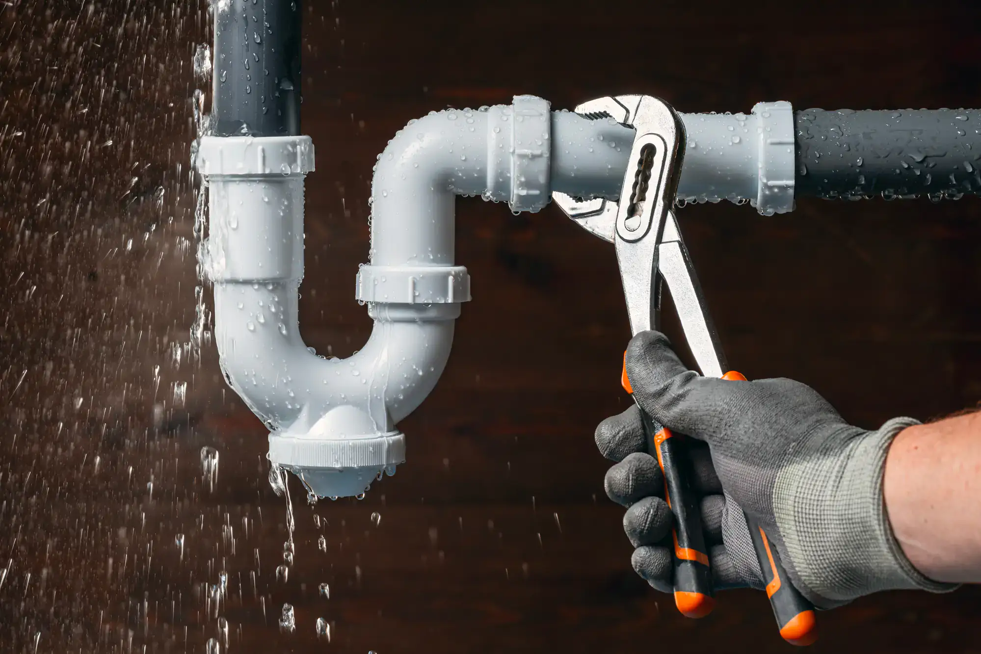A plumber in El Dorado County, CA, wearing gloves, uses a wrench to tighten a leaking white plastic pipe under a sink, with water spraying out from the joint.