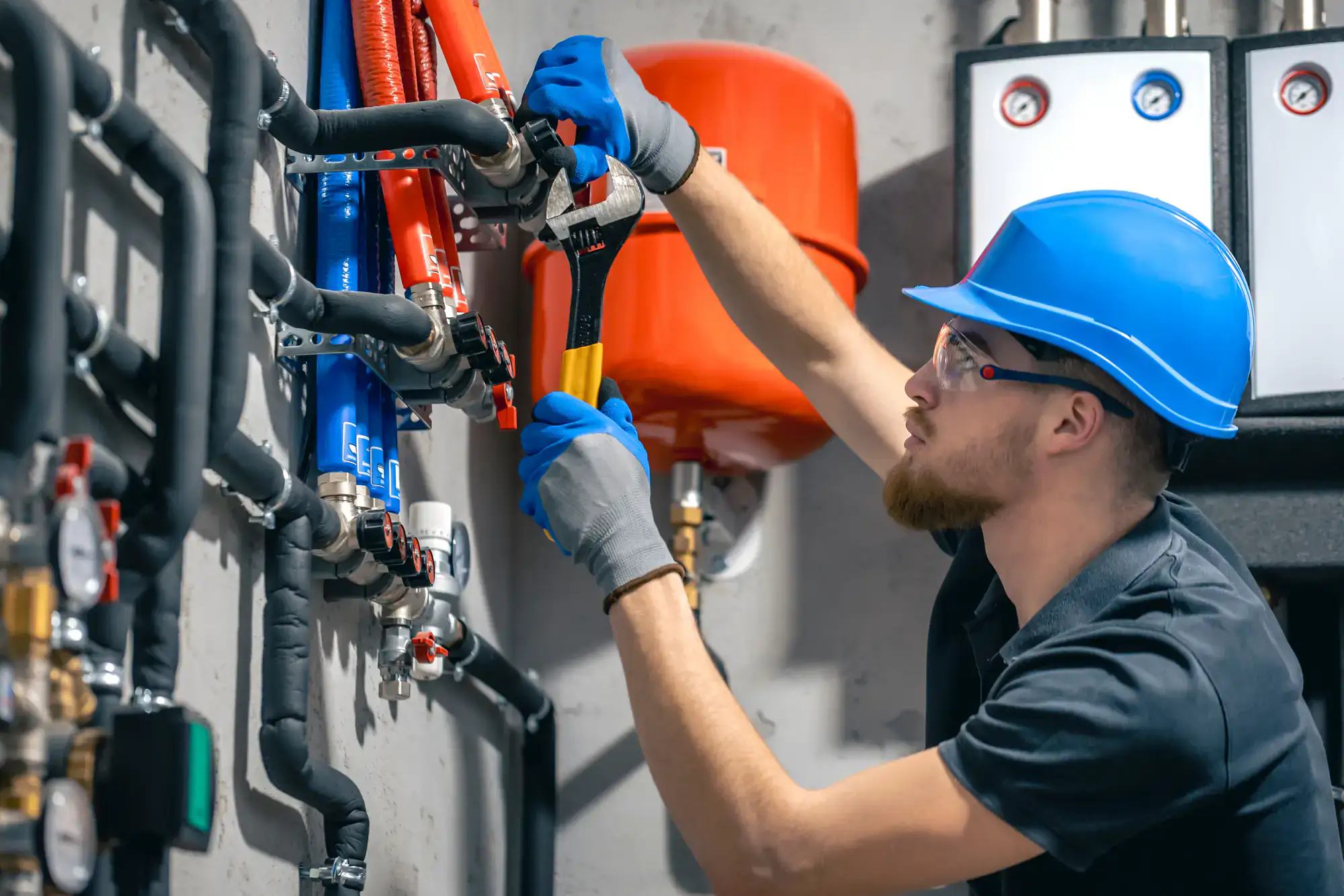 A plumber El Dorado County, wearing a blue hard hat, gloves, and safety glasses, uses a wrench to tighten fittings on pipes and valves in an industrial or mechanical room.