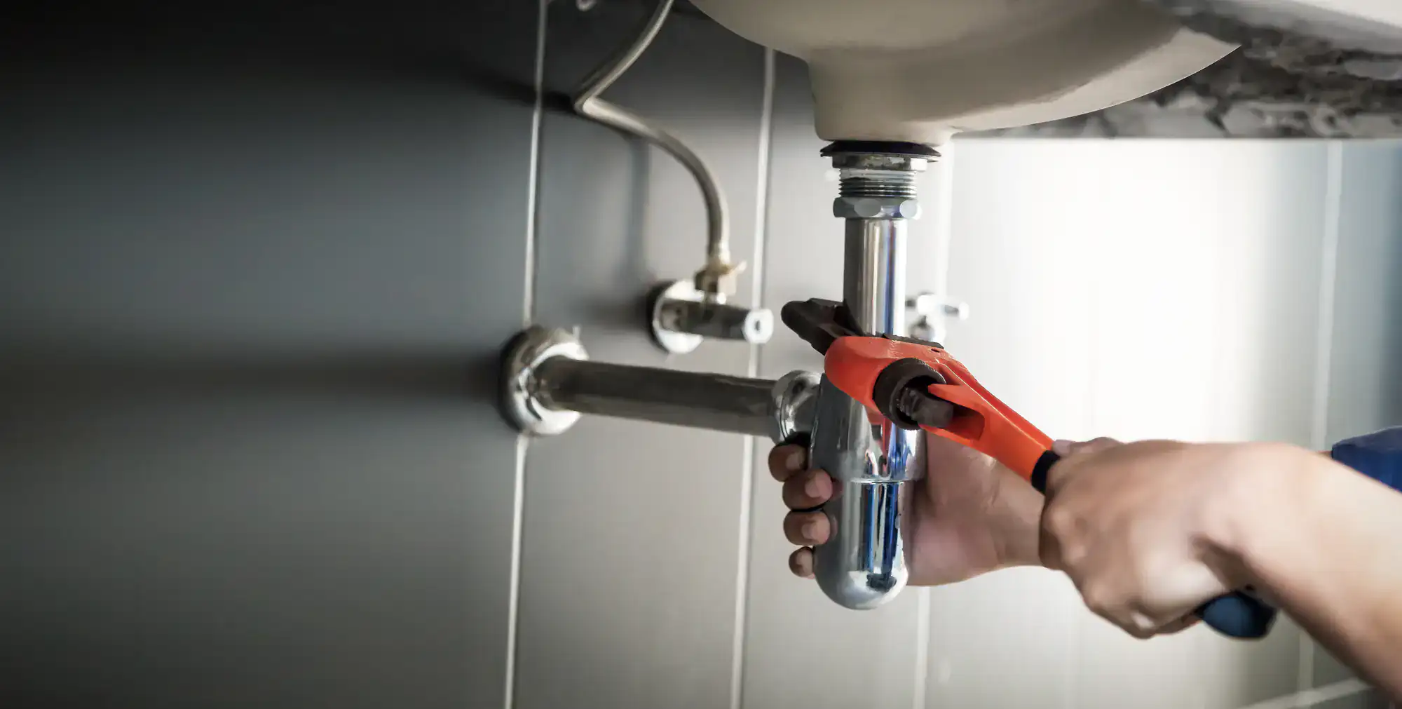 A plumber El Dorado County uses a red pipe wrench to tighten plumbing under a sink, focusing on the metal drainpipe and fittings against a tiled wall.