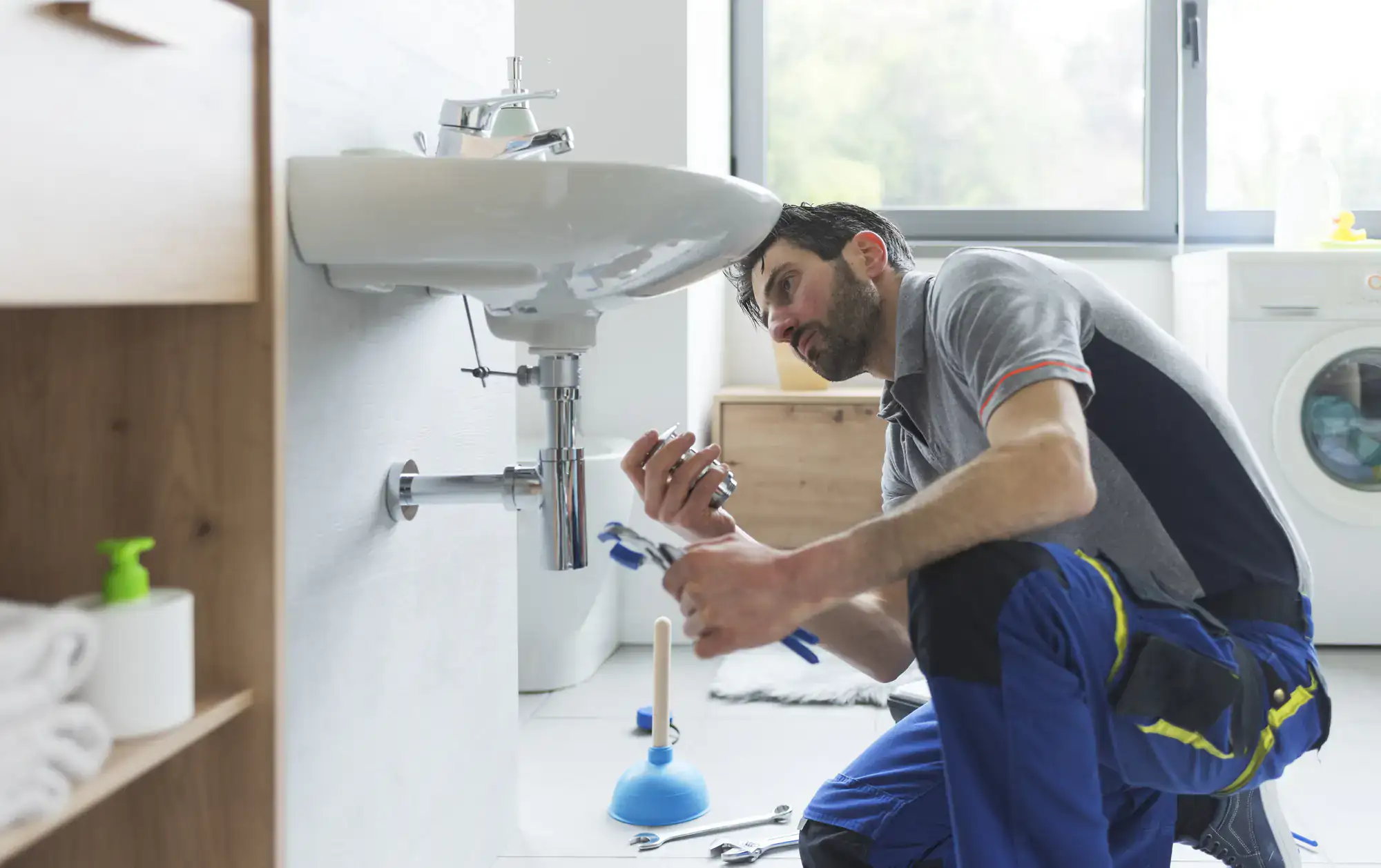 A plumber El Dorado County kneels on a bathroom floor, inspecting and repairing pipes under a wall-mounted sink. Tools and a plunger are nearby, with a washing machine in the background—typical duties for professionals handling water heater replacement El Dorado County.