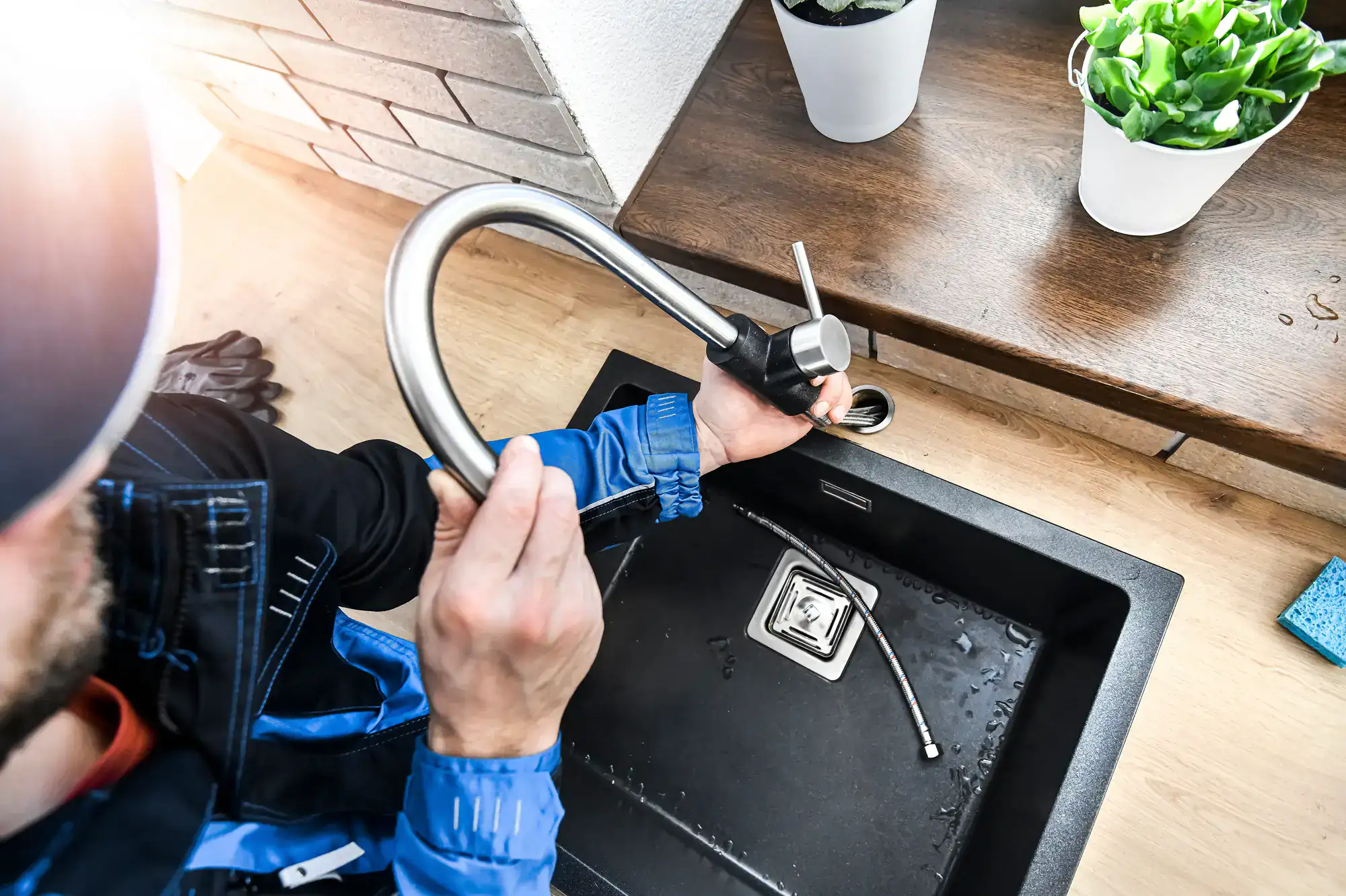 A plumber El Dorado County, CA in a blue uniform installs a new kitchen faucet at a black sink, holding faucet parts while tools and plants are visible on the nearby counter.