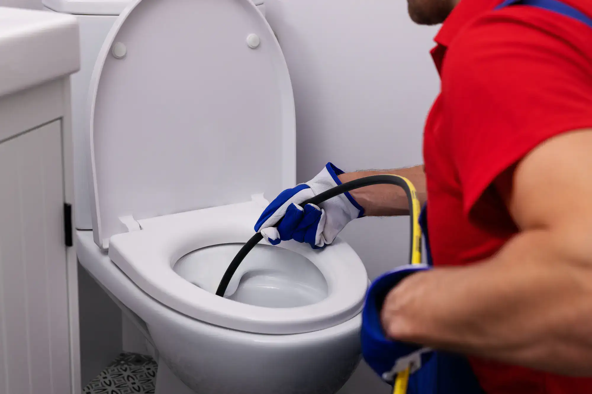 A plumber El Dorado County in a red shirt and blue gloves uses a plumbing snake to unclog a white toilet in a bathroom.
