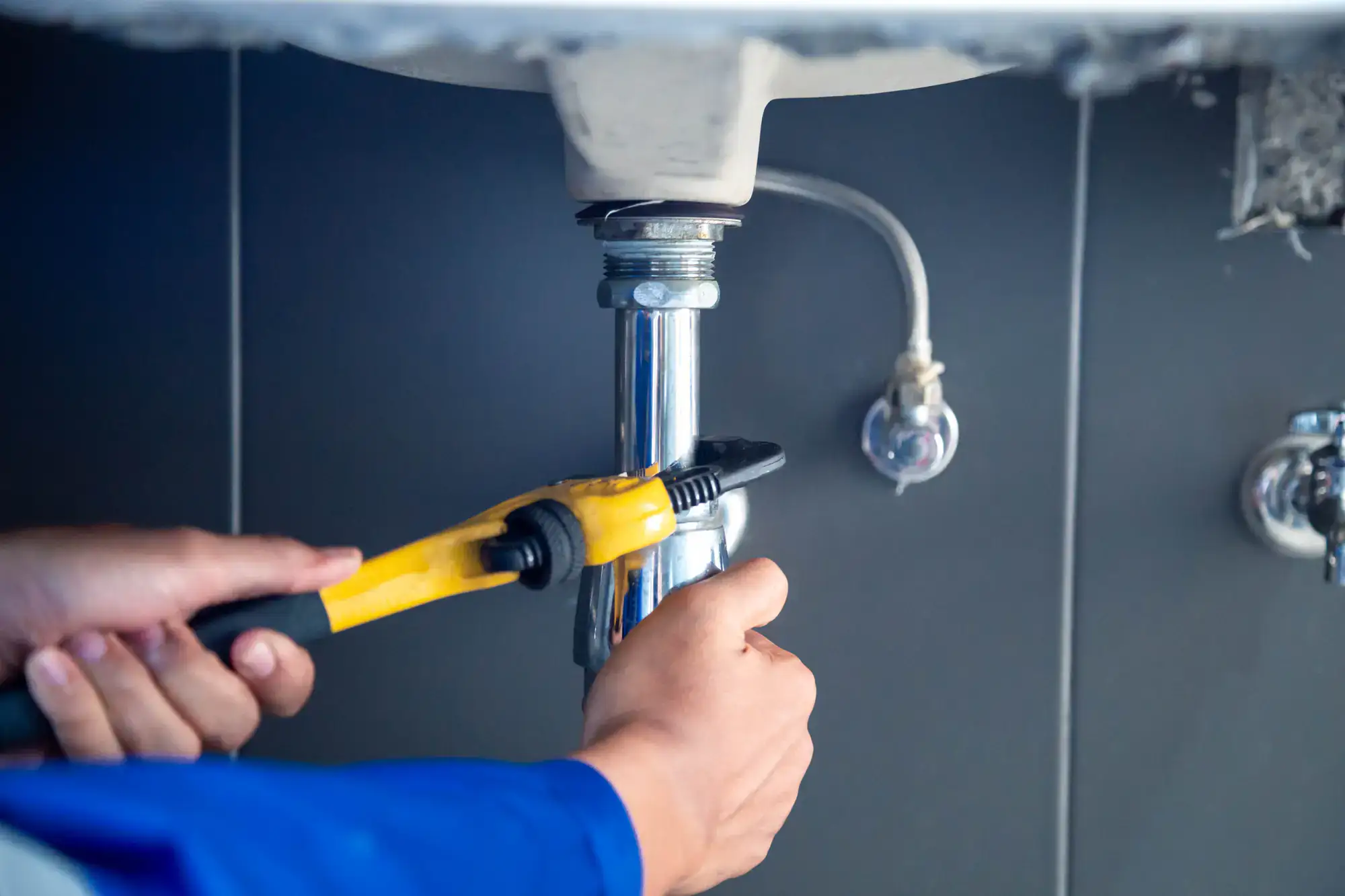 Close-up of a plumber El Dorado County using a yellow pipe wrench to adjust a metal pipe under a sink, set against a gray tiled wall.
