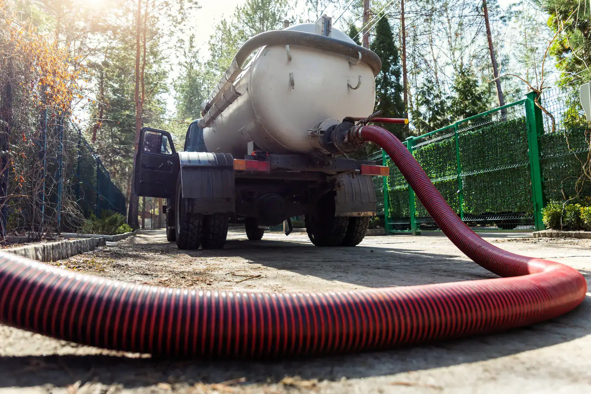 A vacuum truck with a large red hose attached is parked on a paved road near a green fence and trees, possibly supporting a plumber El Dorado County job. The photo is taken from a low angle.