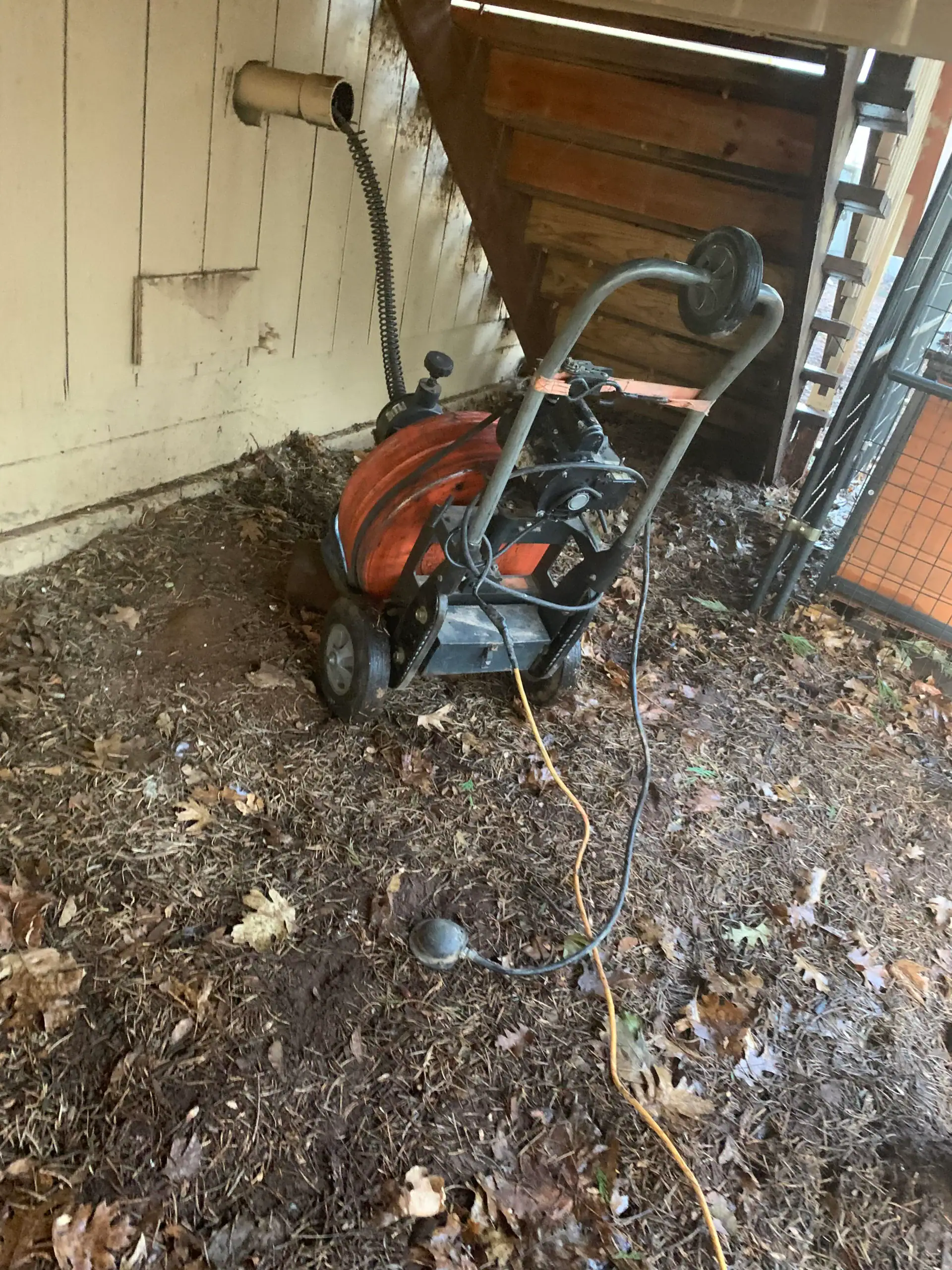 A large drain cleaning machine with an orange cable reel sits on muddy ground under a wooden staircase, next to a wall with pipes and cords—typical tools for a plumber El Dorado County, CA relies on for repairs amid leaves and debris.
