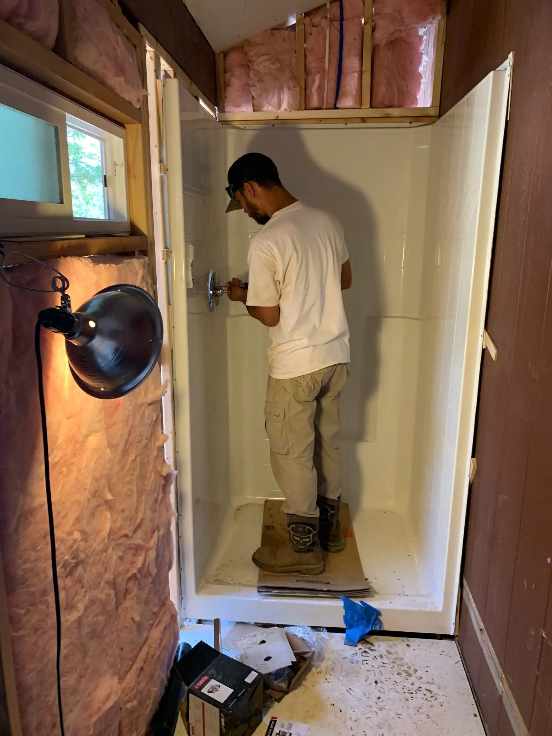 A plumber El Dorado County works inside a white shower stall in a room under construction, surrounded by exposed insulation, tools scattered on the floor, and a work lamp illuminating the scene.