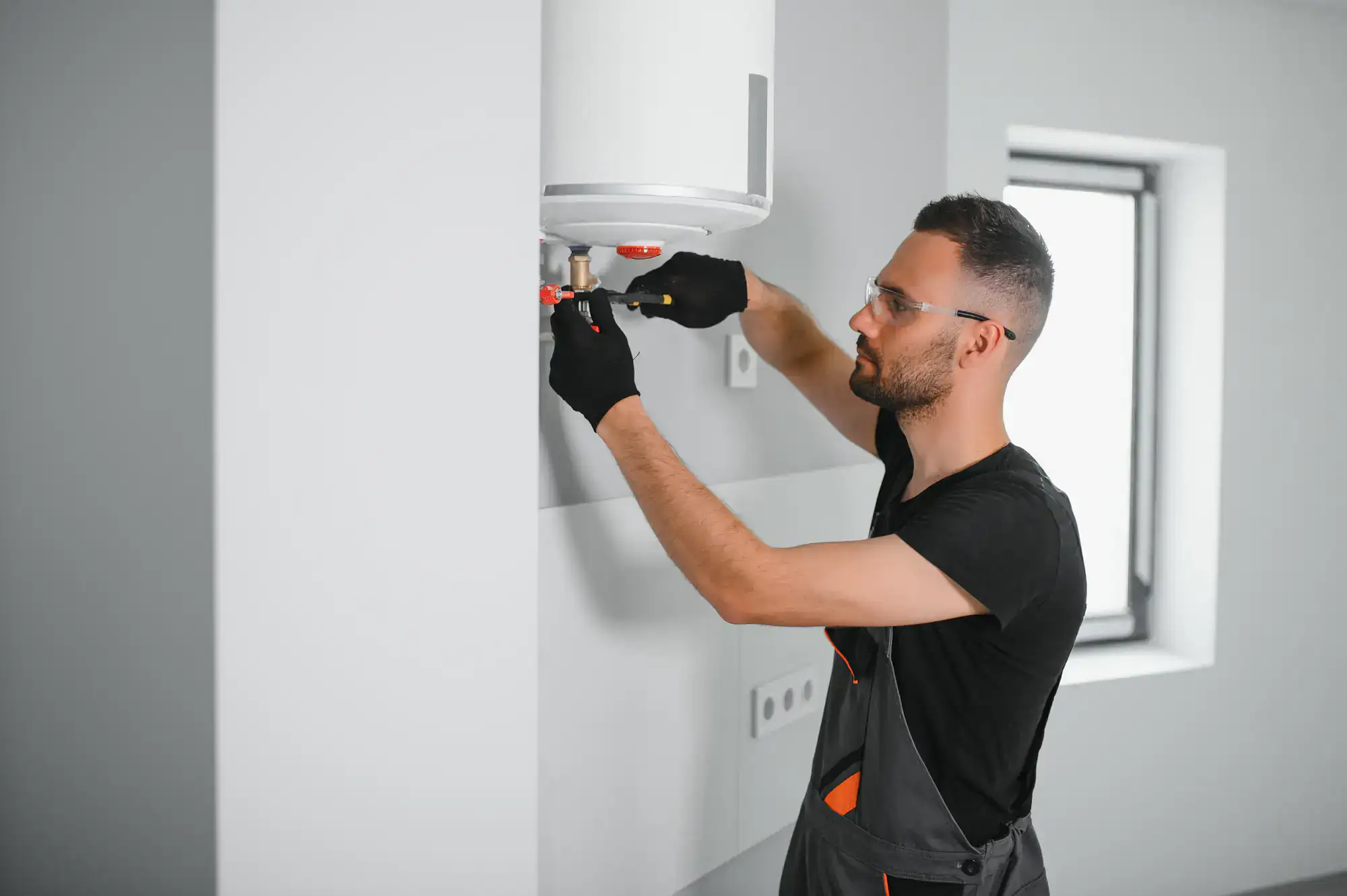 A man wearing gloves and safety glasses, likely a plumber El Dorado County, is using tools to repair or install a water heater mounted on a white wall inside a modern room.
