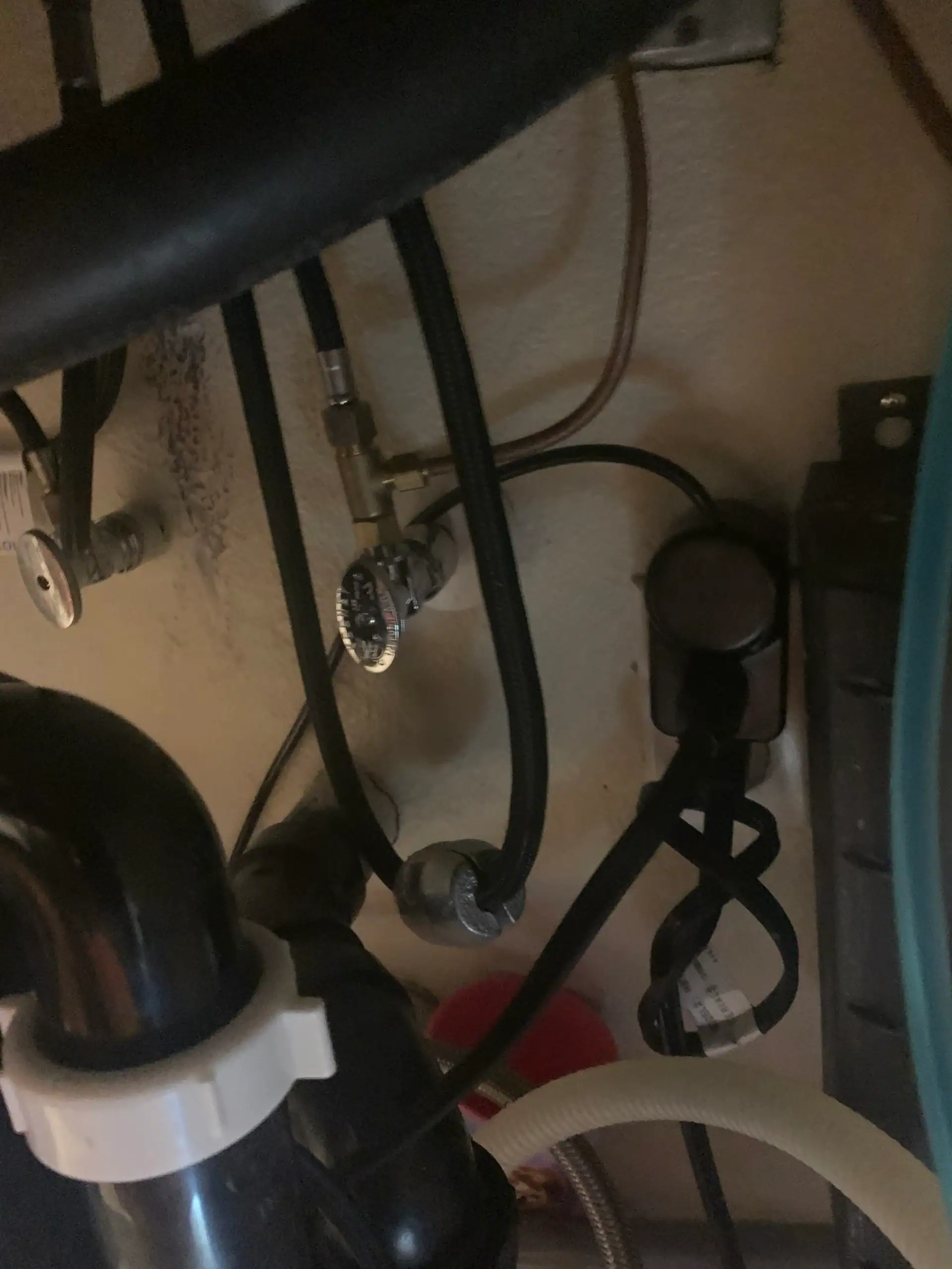 Dimly lit under-sink area in CA, with water pipes, a shutoff valve, black drain pipes, electrical cords, and hoses against a beige wall—ideal for inspection by a plumber El Dorado County. Some details are obscured by low lighting.