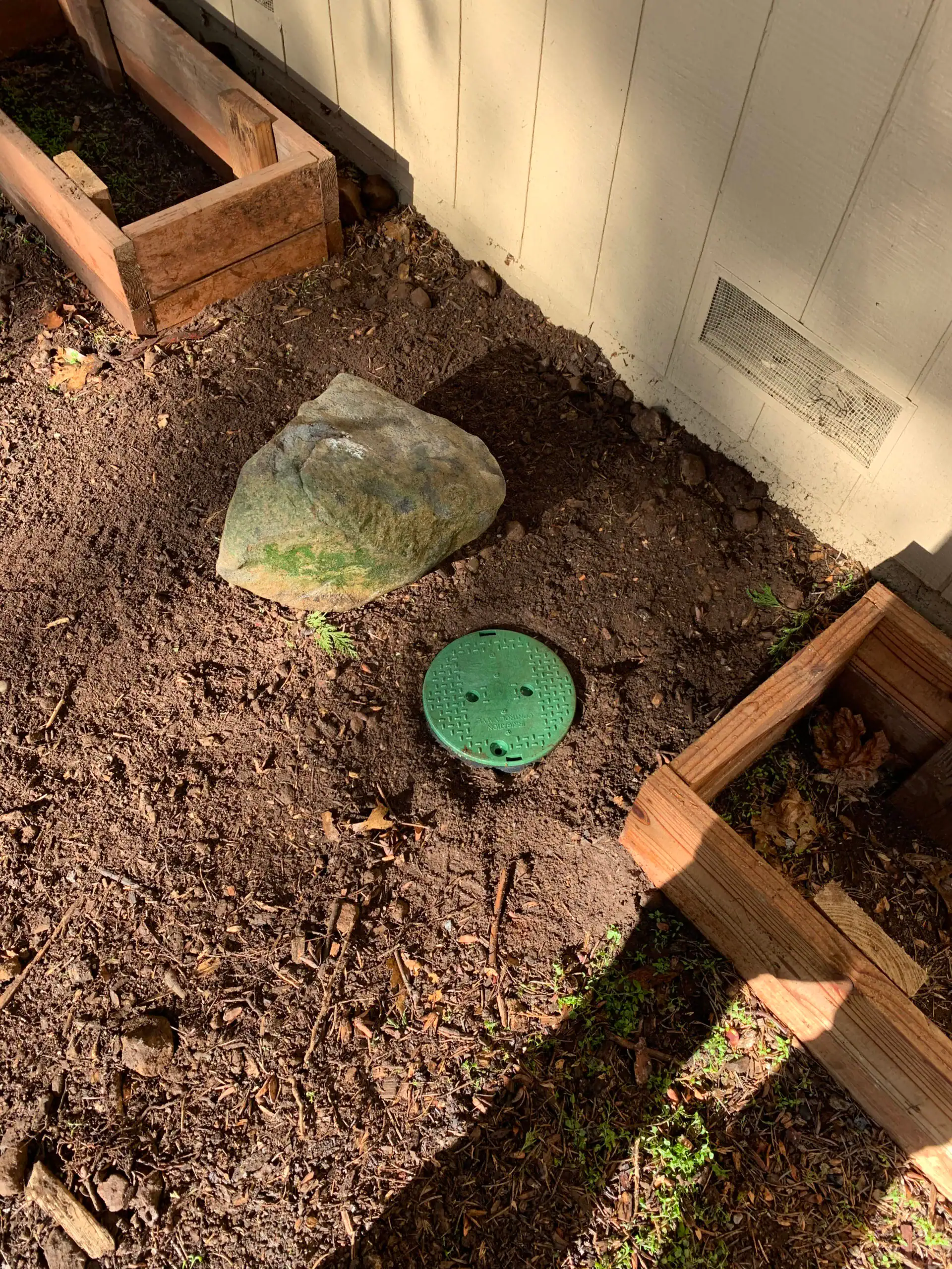 A patch of soil near a house wall in El Dorado County, CA, features a green irrigation valve cover, a large rock, wooden garden bed frames, and sunlight casting shadows—perfect inspiration for your local plumber El Dorado County project.