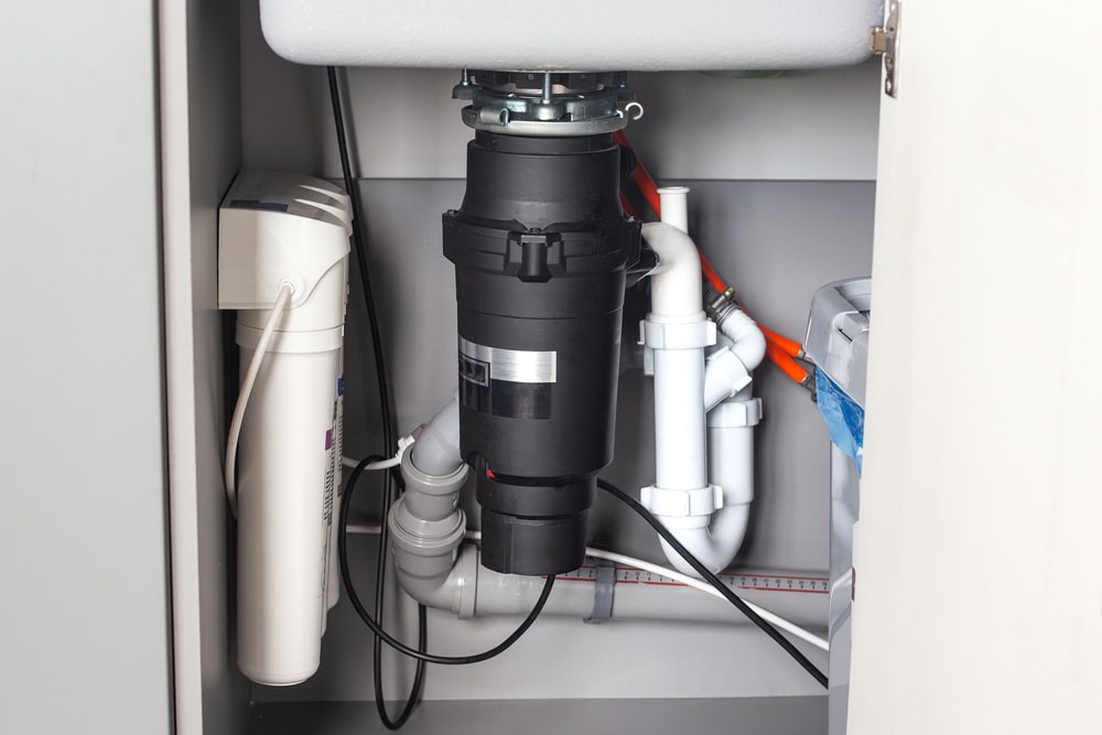 Under-sink view showing a black garbage disposal unit connected to white plumbing pipes, with additional water filter equipment and electrical cords visible inside a cabinet—typical of installations by a plumber El Dorado County, CA.