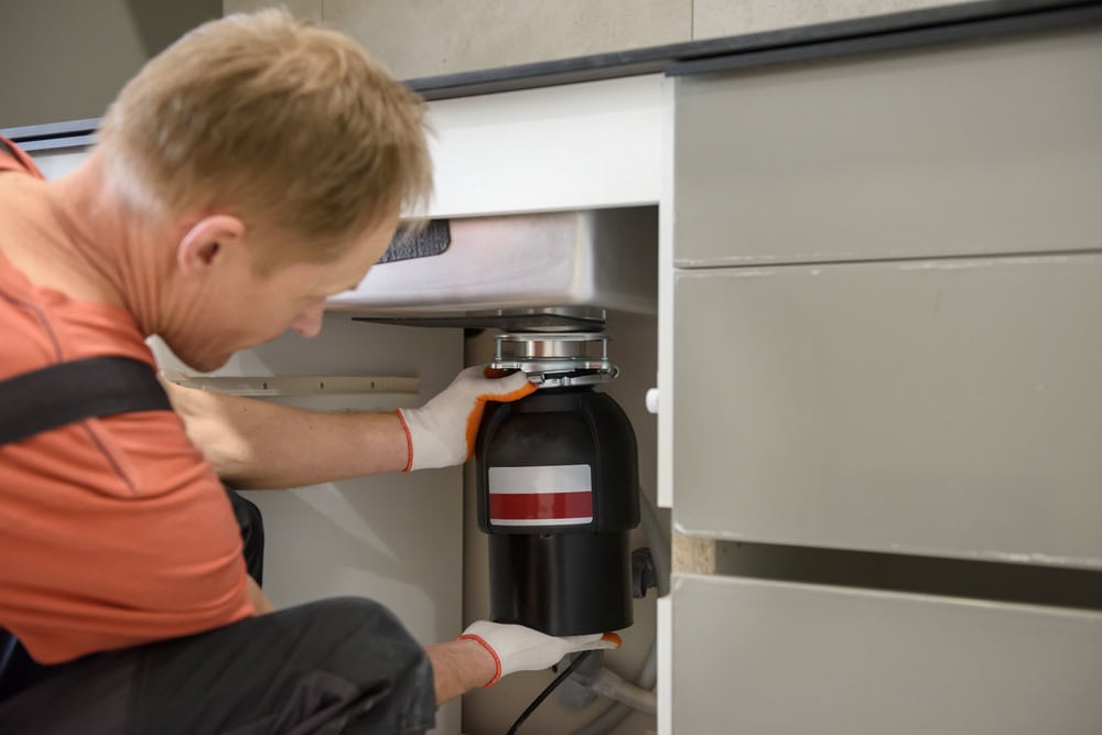 A plumber El Dorado County, CA, wearing gloves installs or repairs a garbage disposal unit under a kitchen sink, with cabinet doors open and tools visible.
