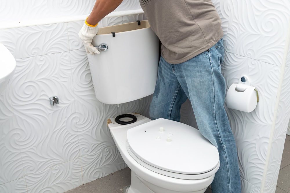 A plumber El Dorado County, wearing gloves and jeans, is installing or repairing a toilet tank in a bathroom with white, textured swirl-patterned walls.