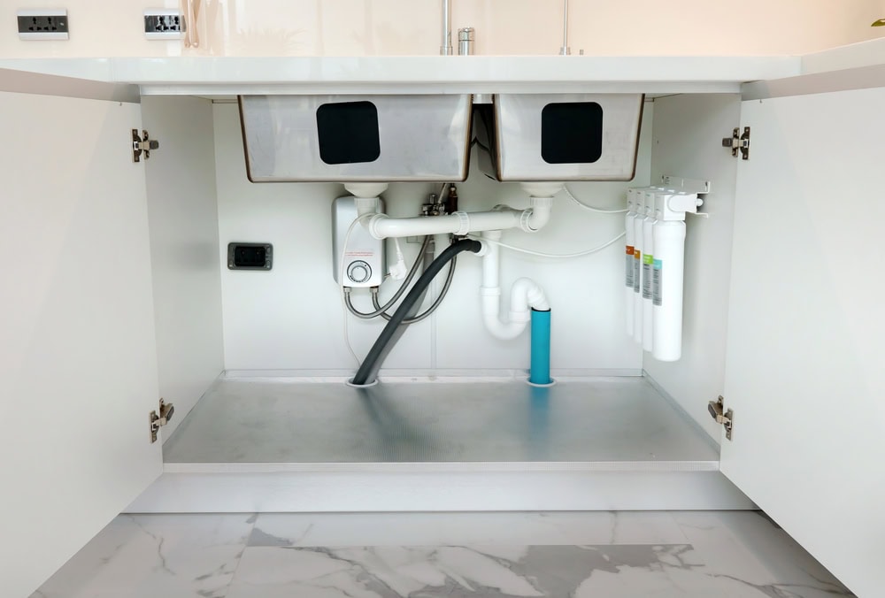 Under-sink view of a kitchen cabinet in El Dorado County, CA, showing plumbing pipes, water filtration system, water heater replacement work, and two stainless steel sink basins above, with cabinet doors open and tiled floor visible.
