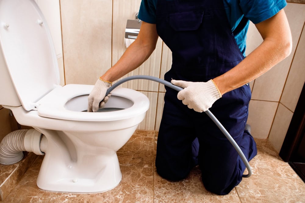 A plumber El Dorado County, CA, wearing gloves and overalls, kneels by a toilet, using a flexible hose to unclog or clean it in a tiled bathroom.