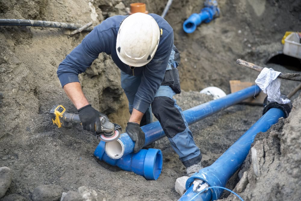 A construction worker wearing a helmet and safety gear uses a power tool to cut a large blue plastic pipe at a busy site, showing the expertise needed for plumber El Dorado County, CA projects like water heater replacement. Dirt and other pipes are visible nearby.