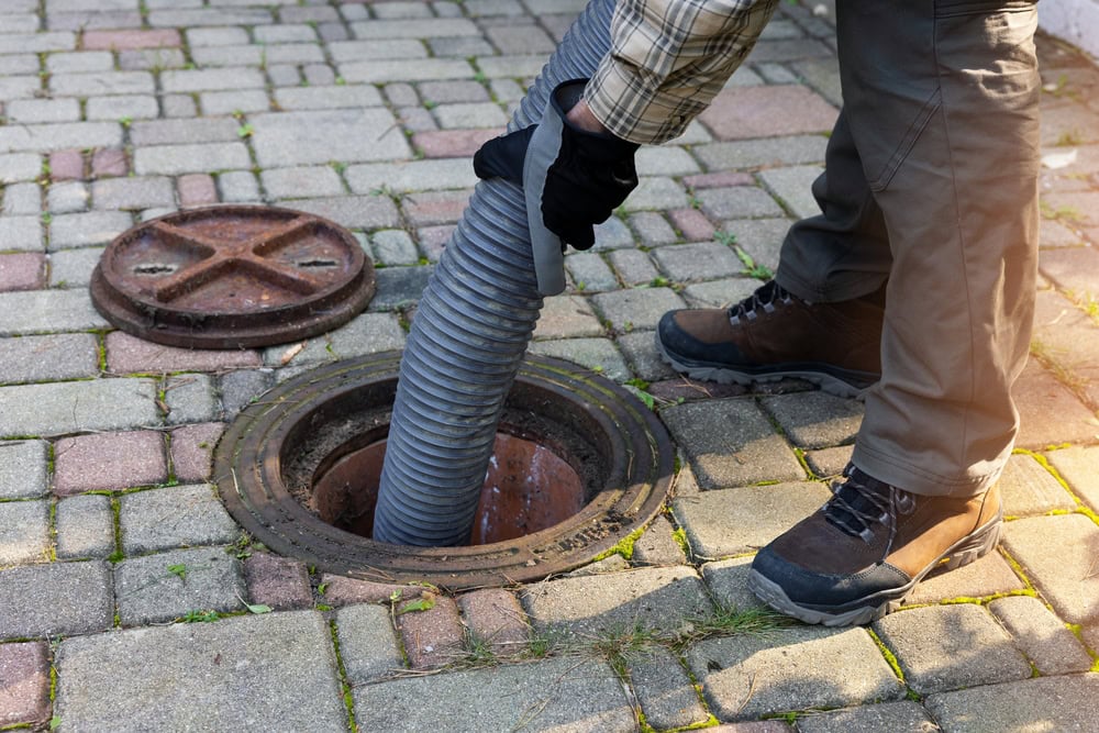 A plumber in El Dorado County, wearing boots and gloves, uses a large hose to clean or empty an outdoor septic tank through an open manhole on a paved surface, with the manhole cover placed nearby.
