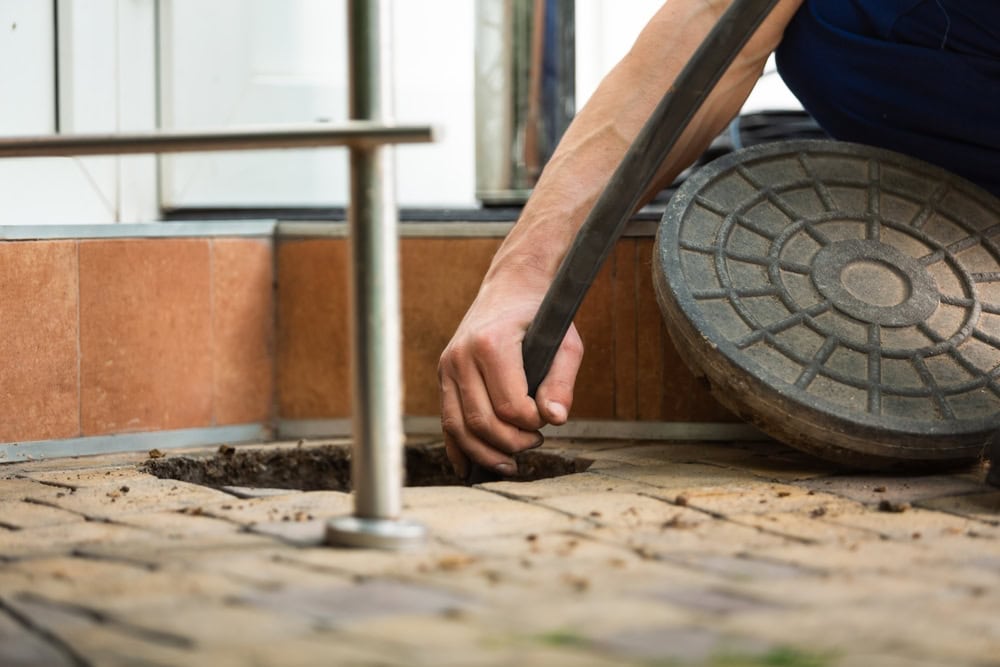 A plumber in El Dorado County lifts a round metal manhole cover and inserts a hose into an outdoor drain on a tiled surface, possibly for cleaning or maintenance work.