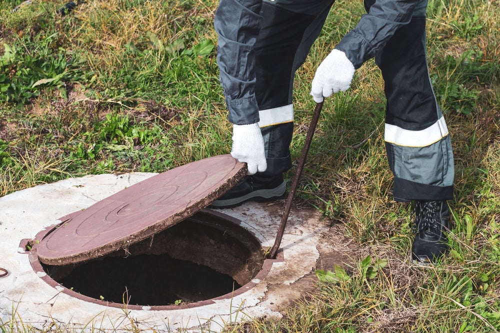 A plumber El Dorado County, CA, wearing protective clothing and white gloves, uses a tool to lift an open manhole cover on a grassy area. The person stands next to the uncovered manhole.