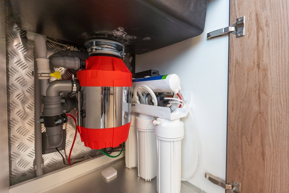 A kitchen sink cabinet interior shows a garbage disposal unit with a red cover, various pipes installed by a plumber El Dorado County, and a white multi-stage water filtration system mounted to the side.