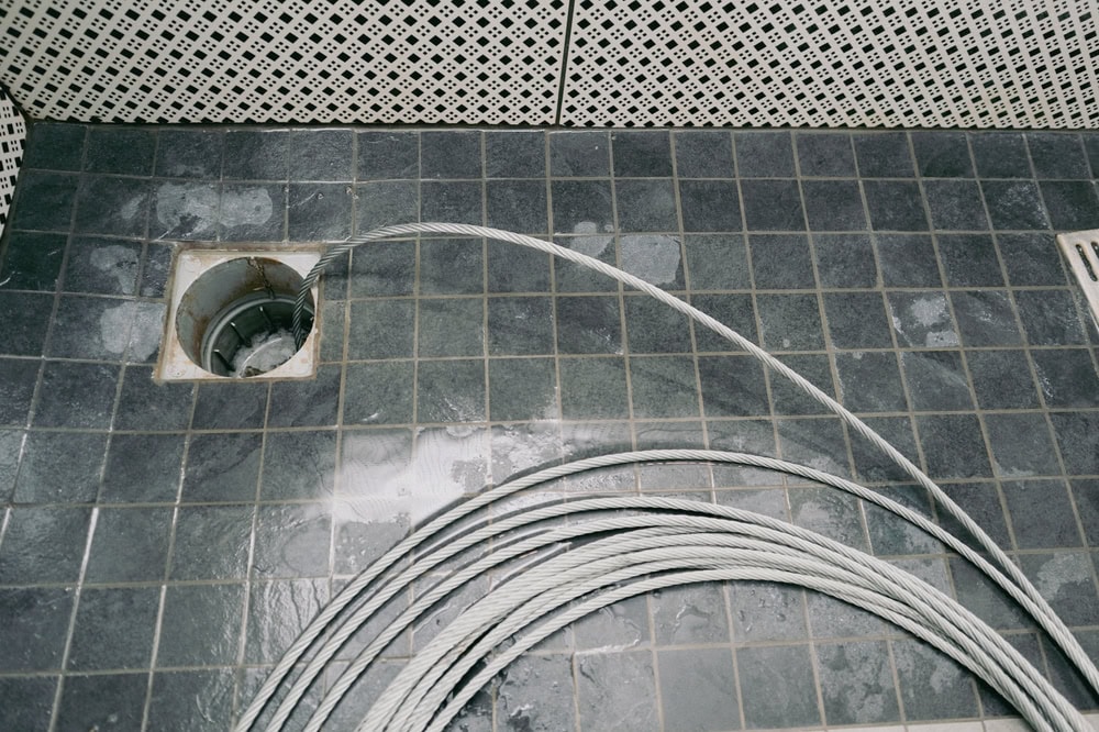 A coil of metal cable lies on a dark tiled floor next to an open drain surrounded by wet patches—just the kind of scene a plumber El Dorado County expert might encounter during water heater replacement or repairs. White patterned tiles line the walls in the background.