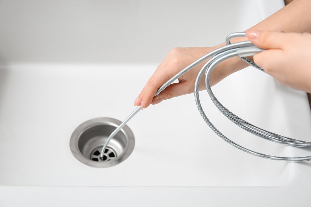 A person’s hands using a plumbing snake tool to unclog a kitchen sink drain in a white sink, demonstrating skills often needed by a plumber El Dorado County, CA residents can trust.