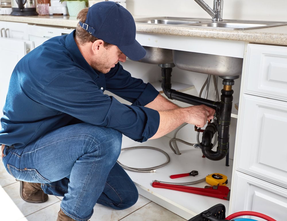 A plumber El Dorado County, CA, wearing a blue shirt and cap kneels under a kitchen sink, repairing pipes with tools nearby—including a wrench, tape measure, and hose.
