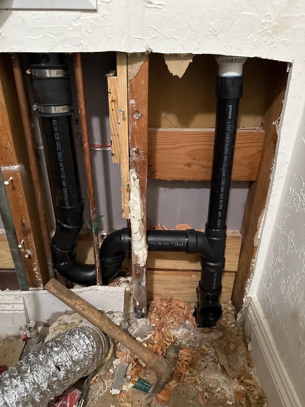 A section of wall is cut open to reveal black PVC plumbing pipes, exposed wooden studs, insulation, and debris on the floor. A hammer and flexible metal duct are also visible near the bottom of the image.