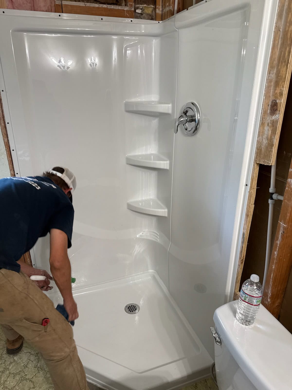 A person wearing a cap cleans a newly installed white shower with built-in shelves; unfinished walls and a water bottle on a nearby toilet are visible, suggesting recent work by a plumber El Dorado County, CA.