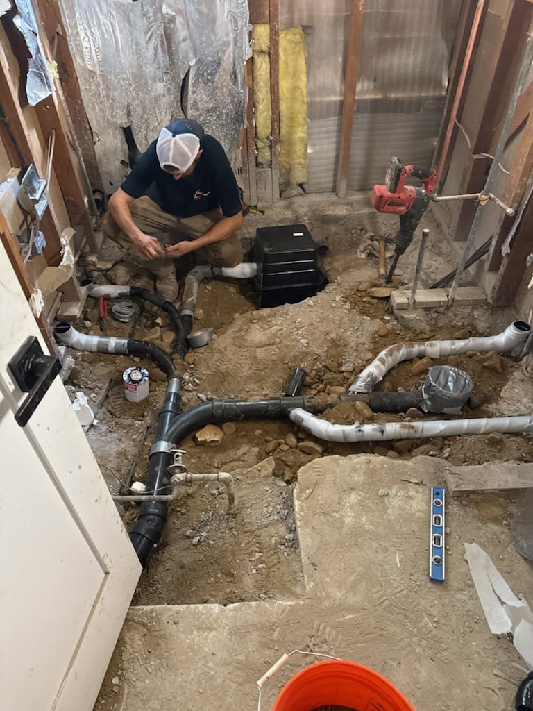 A plumber works on exposed pipes in a bathroom under renovation, surrounded by dirt, tools, and construction materials. Wall studs and insulation are visible, with the floor partially dug up for plumbing installation.