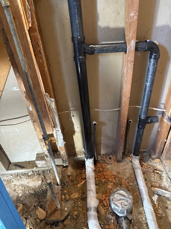 Black PVC plumbing pipes run vertically and horizontally along an exposed wall frame, with some pipes wrapped in white insulation and wood debris scattered on the unfinished ground below.
