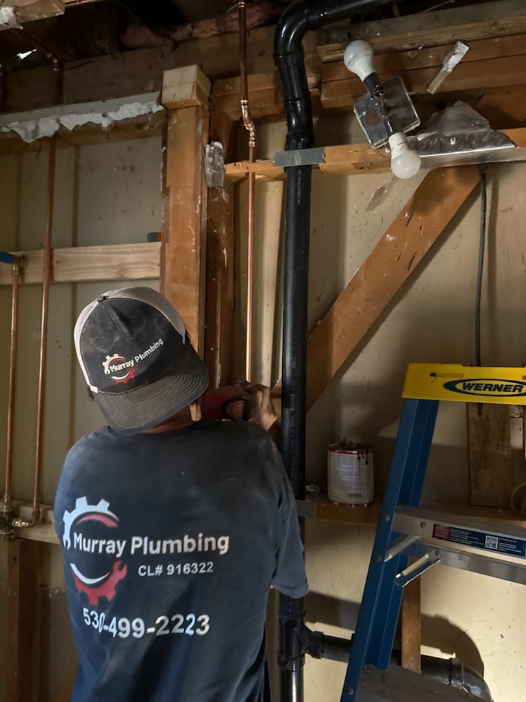 A plumber wearing a Murray Plumbing shirt works on copper pipes in a partially constructed room, with exposed wooden beams, black PVC pipes, a can of paint, and a blue ladder nearby.