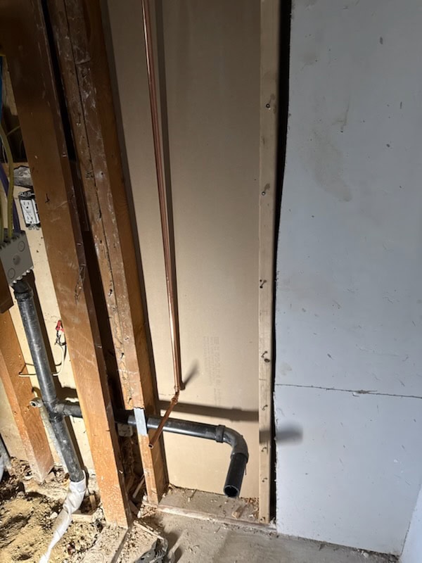 Copper and black PVC pipes are installed inside a partially finished wall with exposed wooden studs and electrical wiring, next to a section of wall with drywall paneling.