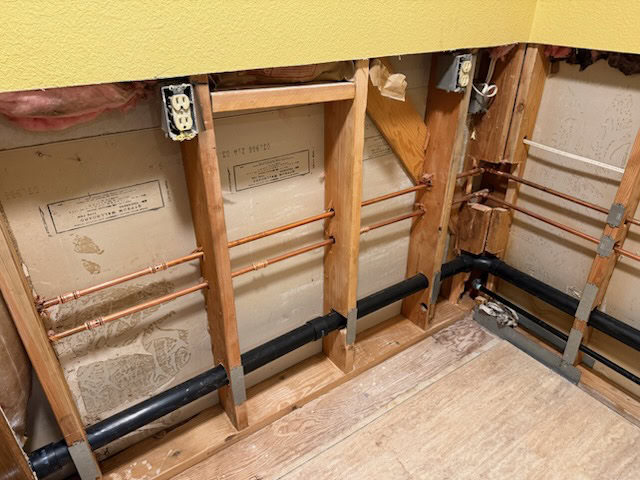 Exposed wall studs with visible copper pipes, black plastic pipes, electrical outlets, and insulation in a room under renovation with a yellow painted wall and plywood floor.