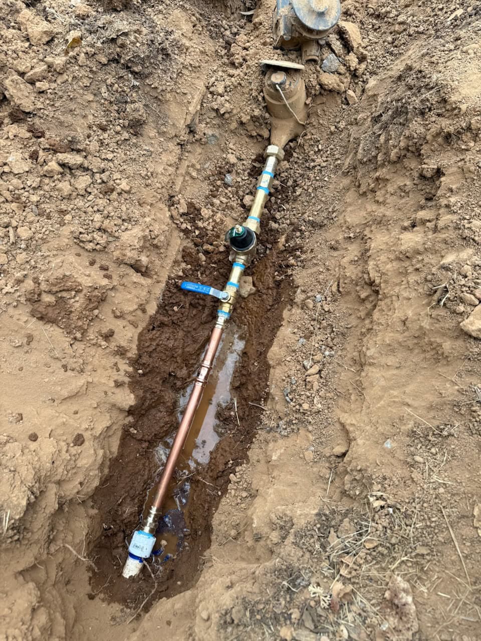 A newly installed water pipe and meter by a plumber in El Dorado County, CA, are exposed in a narrow trench dug into brown soil, with a small amount of water pooled around the pipe near the bottom.