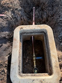A water meter and shut-off valve installed in an underground concrete box, surrounded by soil and mud. Copper pipes are connected to the meter, with a blue-handled valve visible—expertly maintained by a plumber El Dorado County trusts.