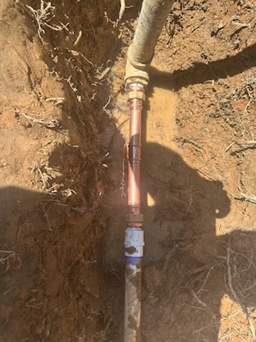 A plumber El Dorado County, CA is fixing a copper pipe in a muddy trench, with fittings joining the sections. The wet ground and exposed roots highlight challenges similar to water heater replacement El Dorado County residents may face.